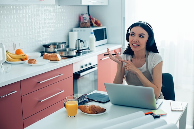 Mujer joven alegre con auriculares sentado frente a la pantalla de un portátil en la cocina y contando algo