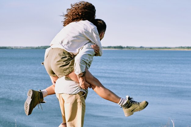 Mujer joven alegre y cariñosa sentada en la espalda de su esposo o novio mientras ambos disfrutan del tiempo junto al mar en un día de verano