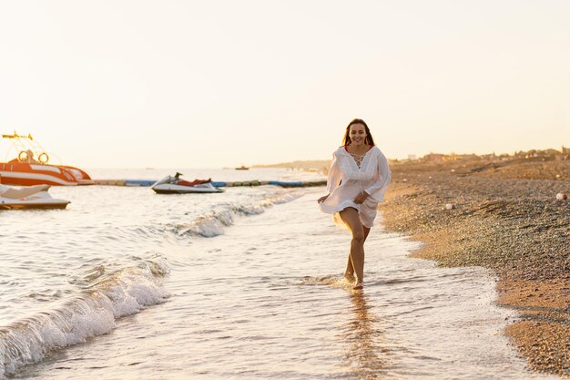 Mujer joven alegre corriendo a lo largo de una playa de arena al atardecer con barcos en el fondo