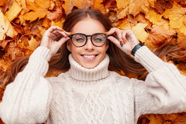 La mujer joven alegre divertida del inconformista en un suéter de punto caliente endereza las gafas de moda. Chica alegre con una sonrisa positiva descansa sobre las hojas de naranja en el parque. Feliz día de otoño.