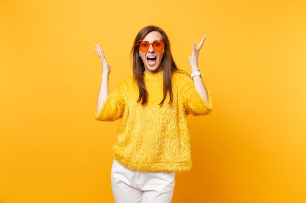 Mujer joven alegre emocionada en suéter de piel, pantalón blanco y gafas de corazón naranja extendiendo las manos aisladas sobre fondo amarillo brillante. Personas sinceras emociones, concepto de estilo de vida. Área de publicidad.