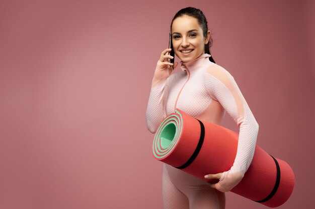 Mujer joven alegre con estera de yoga hablando por teléfono móvil