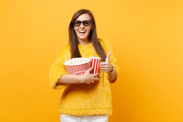 Mujer joven alegre en gafas imax 3d viendo una película, sosteniendo un cubo de palomitas de maíz, una taza de refresco de cola o refresco, mostrando el pulgar hacia arriba aislado sobre fondo amarillo. Personas sinceras emociones en el cine, estilo de vida.