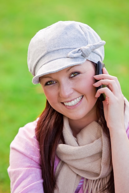 Mujer joven alegre con gorra y bufanda hablando por teléfono en la hierba