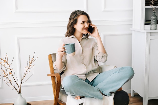 Foto mujer joven alegre hablando por teléfono inteligente relajándose en el sillón con bebida mujer joven feliz hablando