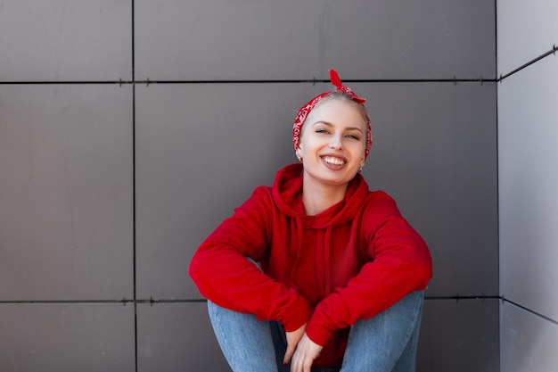 Mujer joven alegre con una hermosa sonrisa en una elegante sudadera roja con un pañuelo de moda en jeans azul está sentada en un día soleado de primavera cerca de un edificio gris