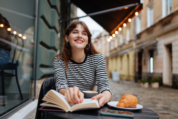 Mujer joven alegre con un libro de papel en la terraza del café