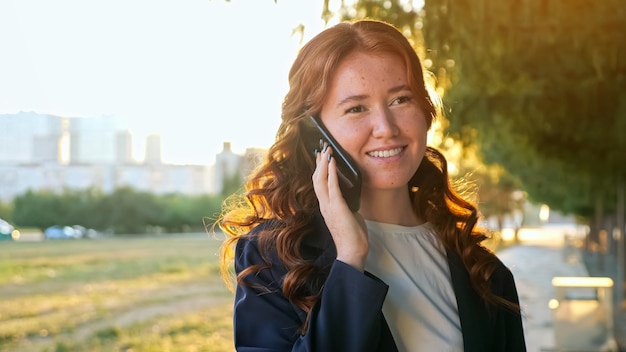 Mujer joven alegre con pecas y cabello rojo que fluye habla sobre el teléfono celular contemporáneo en el parque de la ciudad verde en la vista cercana de la luz del atardecer