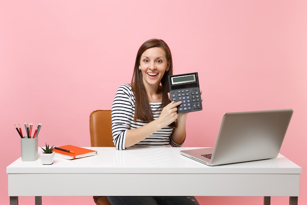 Foto mujer joven alegre que sostiene la calculadora mientras está sentado y trabajando en un proyecto en la oficina con una computadora portátil contemporánea