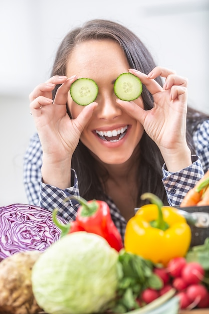 Mujer joven alegre que sostiene rodajas de pepino en la cocina - concepto vegetal y de la salud de la dieta.