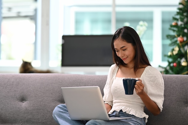 Mujer joven alegre que usa la computadora portátil viendo videos divertidos y tomando café mientras está sentado en el sofá en la sala de estar.