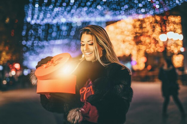 Mujer joven alegre con regalo rojo divirtiéndose en la calle de la ciudad en Navidad.