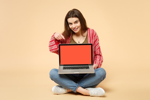 Mujer joven alegre en ropa casual sentada, sosteniendo la computadora portátil con pantalla vacía en blanco aislada sobre fondo beige pastel. Personas sinceras emociones, concepto de estilo de vida. Simulacros de espacio de copia.