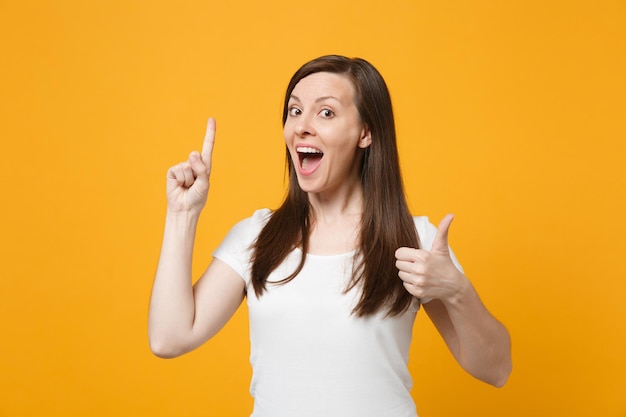 Mujer joven alegre con ropa informal blanca que muestra el pulgar hacia arriba, sosteniendo el dedo índice con una gran idea nueva aislada en un fondo naranja amarillo brillante. Concepto de estilo de vida de las personas. Simulacros de espacio de copia.