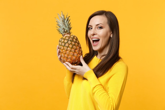 Mujer joven alegre con ropa informal que mantiene la boca abierta, sosteniendo fruta fresca de piña madura aislada en un fondo amarillo anaranjado. Estilo de vida vivo de la gente, concepto de vacaciones relajantes. Simulacros de espacio de copia