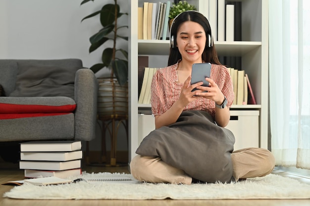 Mujer joven alegre con ropa informal usando un teléfono inteligente y escuchando música o podcast en auriculares inalámbricos