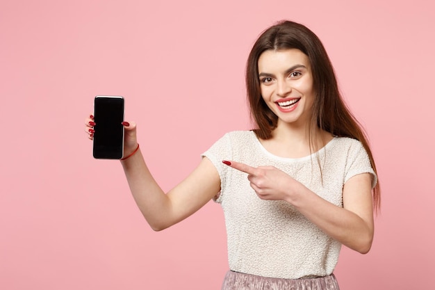 Mujer joven alegre con ropa ligera informal posando aislada en un fondo rosa pastel. Concepto de estilo de vida de las personas. Simulacros de espacio de copia. Señalar con el dedo índice en el teléfono móvil con pantalla vacía en blanco.