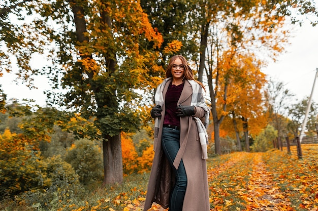 Mujer joven alegre en ropa de otoño cálido de moda corre a lo largo de un sendero en un parque con árboles con hojas de color amarillo anaranjado. Chica alegre con una sonrisa positiva disfruta del fin de semana.