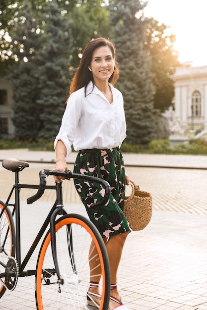 Mujer joven alegre con ropa de verano caminando por una calle de la ciudad con bicicleta