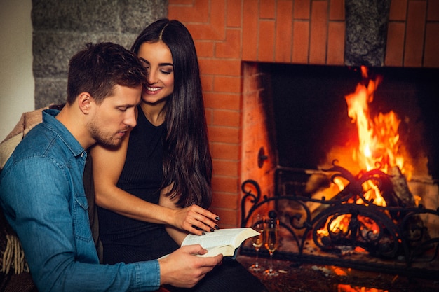 La mujer joven alegre se sienta con el novio y lo mira. Ella sonríe. Guy leyó el libro. El esta concentrado. Pareja sentada en la chimenea.