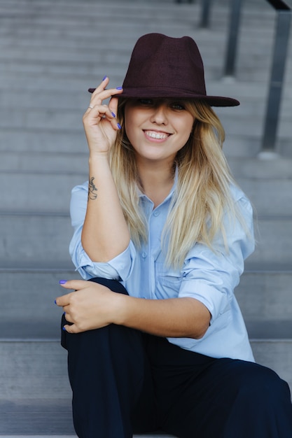 Mujer joven alegre con sombrero elegante, camisa azul y pantalón negro, sentada en los escalones cerca del edificio moderno. Concepto de moda y estilo.