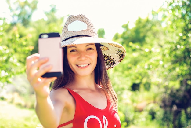 Mujer joven alegre en el sombrero que toma el selfie con el teléfono móvil en centro turístico de verano