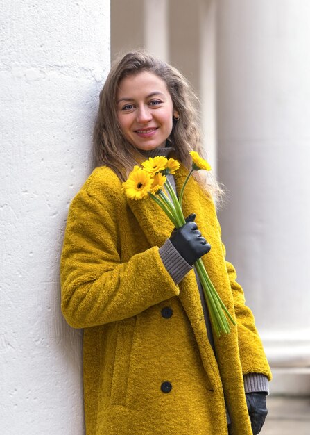 Una mujer joven y alegre con una sonrisa en un abrigo amarillo se encuentra cerca de las columnas con flores amarillas en primer plano