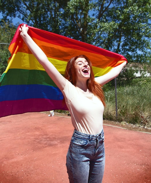 Mujer joven alegre sosteniendo una bandera arco iris en la cancha de baloncesto contra los árboles