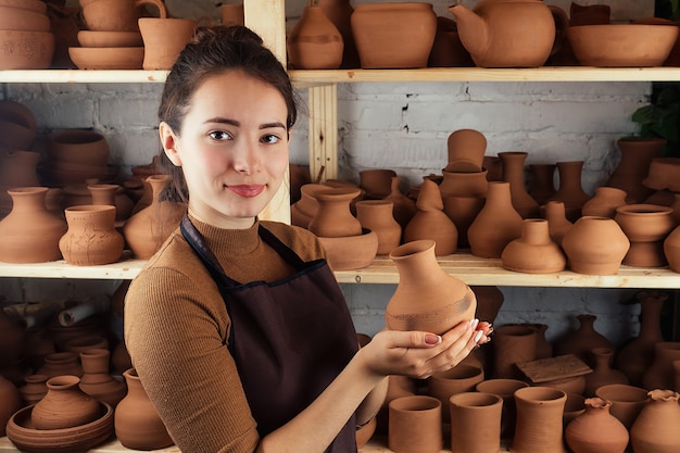 Una mujer joven y alegre sosteniendo un jarrón de barro. El alfarero trabaja en un taller de alfarería con arcilla. el concepto de maestría en cerámica y creatividad.