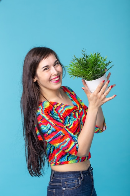 Mujer joven alegre sostiene una maceta con una planta posando contra una pared azul. Concepto de jardinería y accesorios de interior.