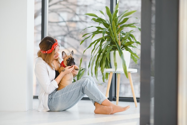 Mujer joven alegre con su gran cachorro con nariz negra y riendo. Retrato interior de niña sonriente posando con bulldog francés