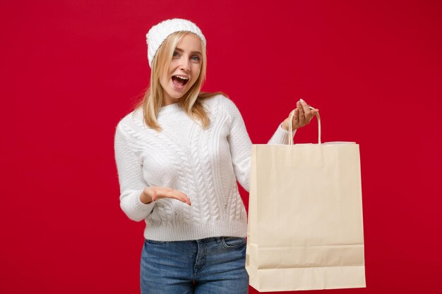 Mujer joven alegre en suéter blanco, sombrero aislado sobre fondo rojo. Estilo de vida de moda saludable, concepto de temporada fría. Simulacros de espacio de copia. Apuntando con la mano en la bolsa del paquete con compras después de comprar.