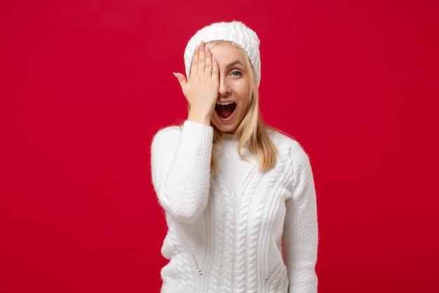 Foto mujer joven alegre en suéter blanco, sombrero aislado sobre fondo rojo, retrato de estudio. estilo de vida de moda saludable, concepto de temporada fría. simulacros de espacio de copia. cubrirse la cara con la mano, manteniendo la boca abierta.