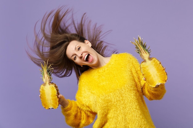 Mujer joven alegre en suéter de piel con cabello que fluye sostiene mitades de fruta de piña madura fresca aislada sobre fondo violeta pastel. Estilo de vida vivo de la gente, concepto de vacaciones relajantes. Simulacros de espacio de copia.