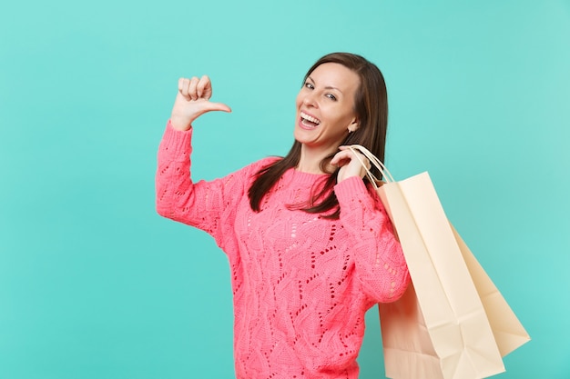 Mujer joven alegre en suéter de punto rosa apuntando con el pulgar sobre sí misma mantenga en la mano paquetes bolsas con compras después de ir de compras aisladas sobre fondo azul. Concepto de estilo de vida de personas. Simulacros de espacio de copia.