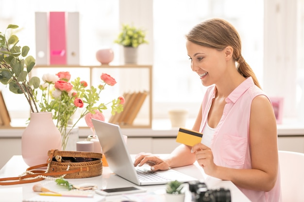 Mujer joven alegre con tarjeta de plástico mirando la pantalla del portátil mientras ingresa datos personales para pagar el pedido en la tienda online