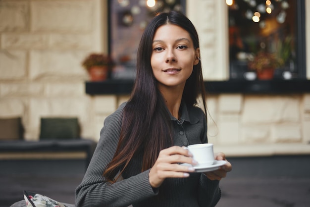 Mujer joven alegre con una taza de café sentado en el café sonriendo al aire libre