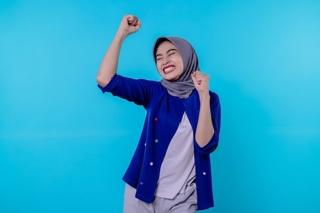 La mujer joven alegre tiene una expresión positiva, aprieta los puños, tiene una mirada llena de alegría, está en alto espíritu, usa hijab, aislado sobre una pared azul