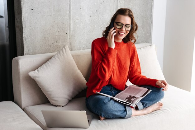 Mujer joven alegre vestida casualmente en auriculares sentado en un sofá en casa, estudiando con ordenador portátil, sosteniendo el teléfono móvil