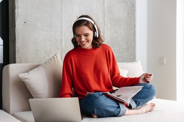 Mujer joven alegre vestida casualmente en auriculares sentado en un sofá en casa, estudiando con ordenador portátil