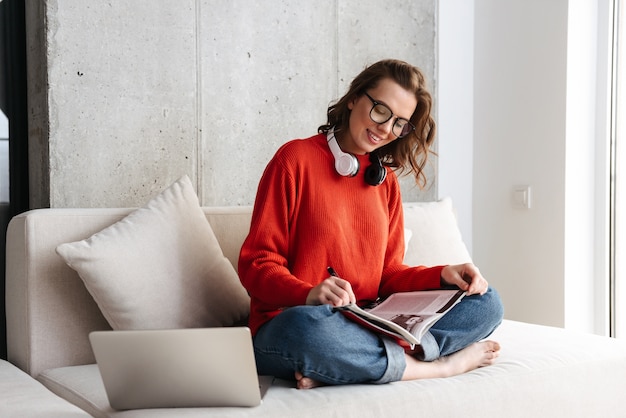 Mujer joven alegre vestida casualmente en auriculares sentado en un sofá en casa, estudiando con ordenador portátil