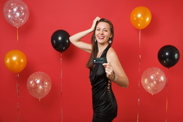 Mujer joven alegre en vestidito negro celebrando, sosteniendo la tarjeta de crédito, poniendo la mano en la cabeza en globos de aire de fondo rojo brillante. Concepto de fiesta de día de fiesta de maqueta de cumpleaños de feliz año nuevo de la mujer.