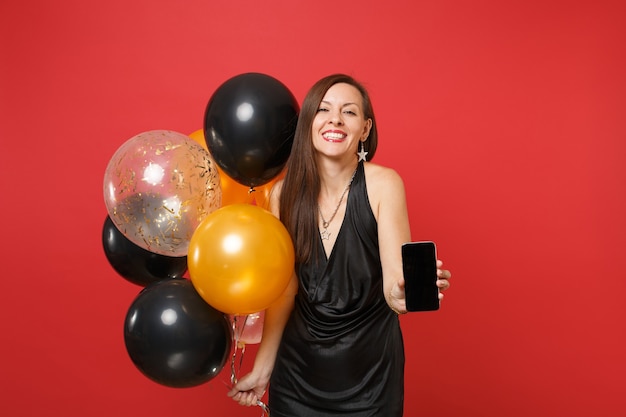 Mujer joven alegre en vestidito negro celebrando, sosteniendo el teléfono móvil con pantalla vacía en blanco negro, globos de aire aislados sobre fondo rojo. Concepto de fiesta de vacaciones de maqueta de cumpleaños de feliz año nuevo.