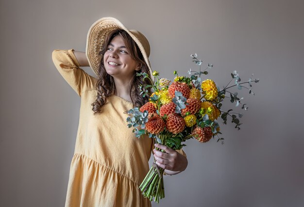 Mujer joven alegre con un vestido amarillo y un sombrero con un ramo de crisantemos brillantes sobre un fondo gris.