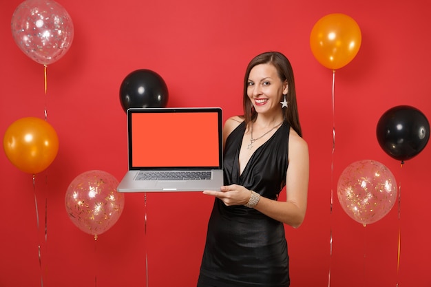 Mujer joven alegre en vestido negro celebrando, sosteniendo la computadora portátil con pantalla vacía en blanco sobre fondo rojo brillante globos de aire. Feliz año nuevo, concepto de fiesta de vacaciones de maqueta de cumpleaños.