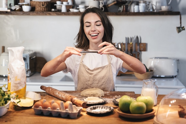 Mujer joven alegre vistiendo delantal preparando la masa para un pastel de manzana en la cocina de casa, haciendo una foto