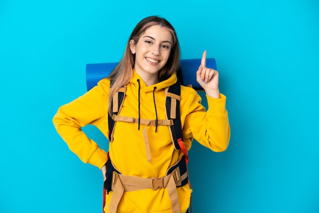 Mujer joven alpinista con una gran mochila aislada en la pared azul mostrando y levantando un dedo en señal de lo mejor