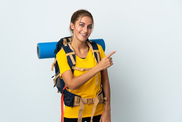 Mujer joven alpinista con una gran mochila aislada en la pared blanca apuntando hacia el lado para presentar un producto