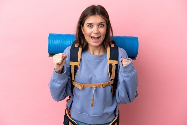 Mujer joven alpinista con una gran mochila aislada sobre fondo rosa celebrando una victoria en la posición ganadora