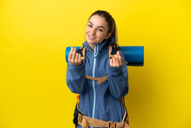 Mujer joven alpinista con una gran mochila sobre fondo amarillo aislado haciendo gesto de dinero